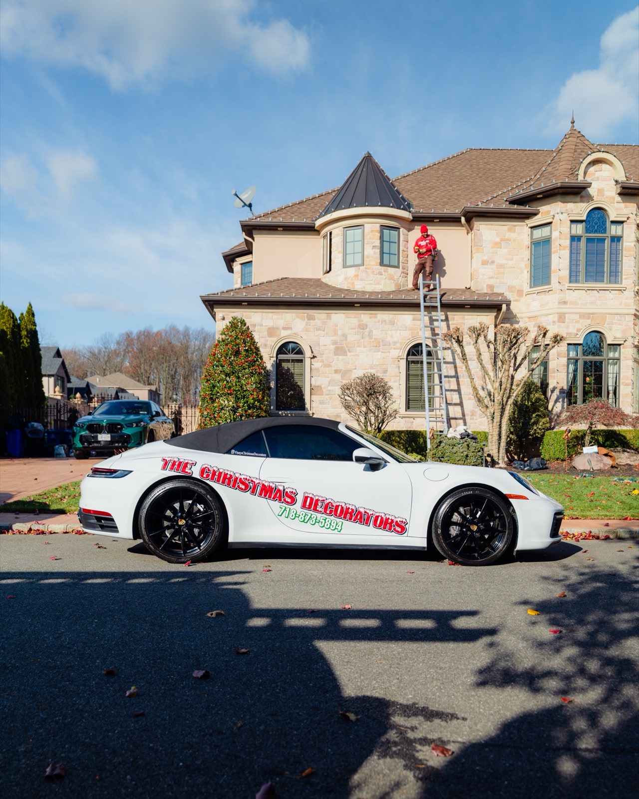 Crew on ladders installing lights on a stone mansion by day, branded Porsche in the driveway
